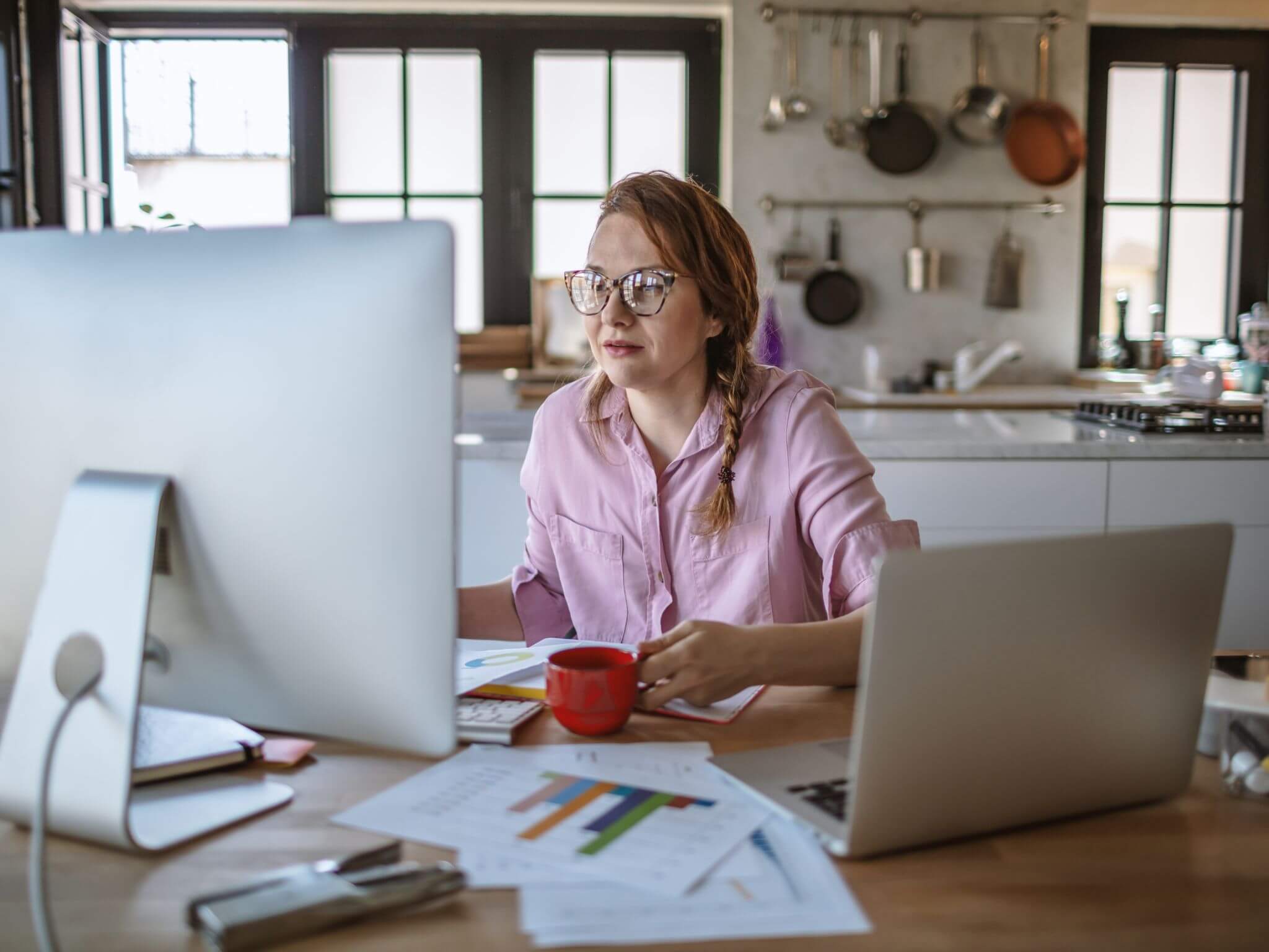 A lady wearing a plait and glasses sitting at a table with a mug of tea reading her computer screen.