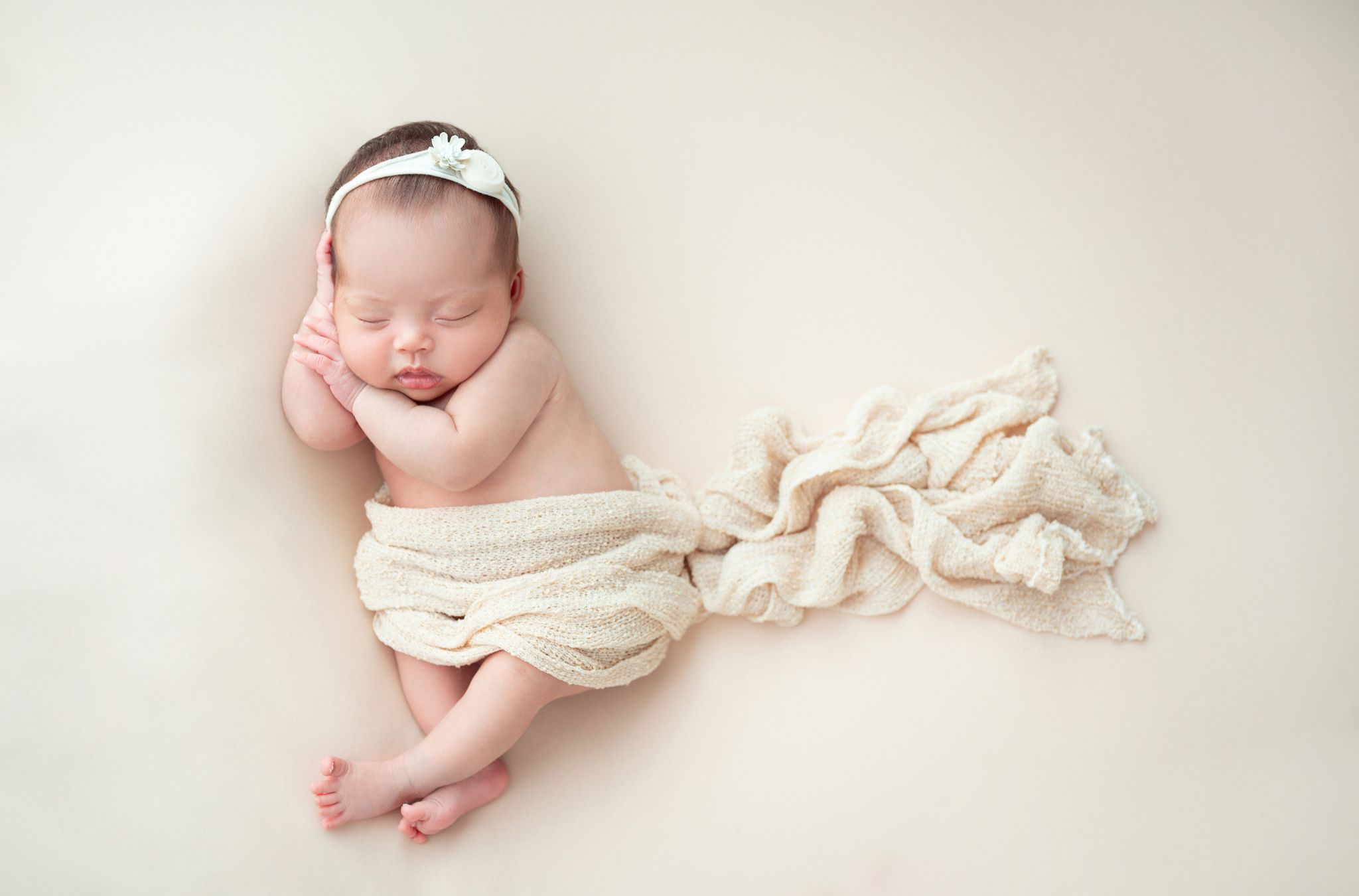 A newborn baby girl sleeping with a cream blanket and a pretty white cloth headband.