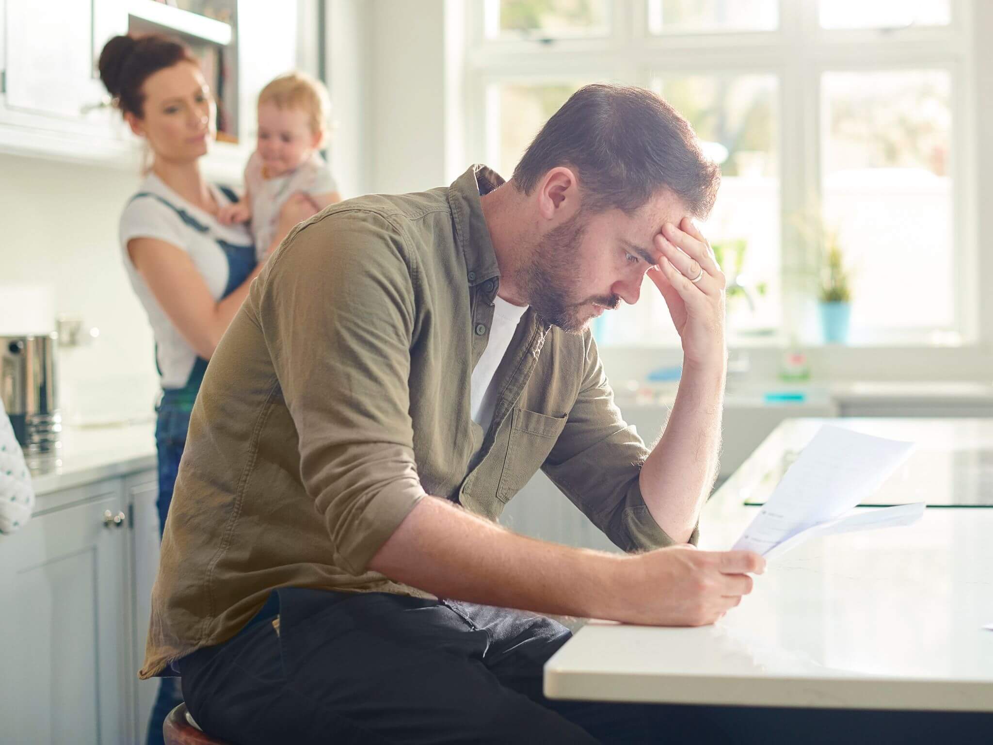 A young father sitting at a table going through bills. His wife and young baby are in the background.