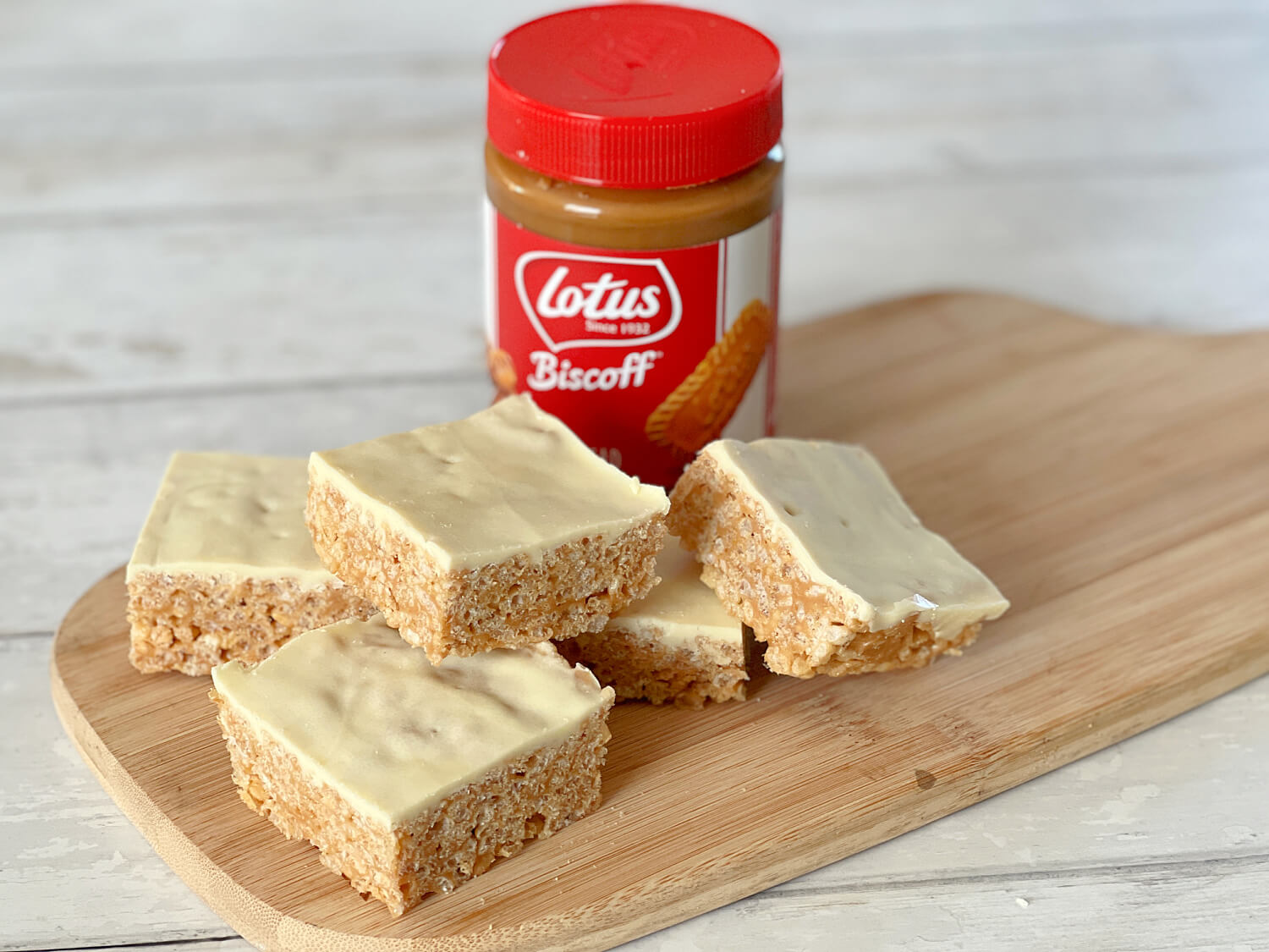 Square Biscoff rice crispy cakes with white chocolate topping. Displayed on a wooden serving board next. to a jar of Biscoff spread.