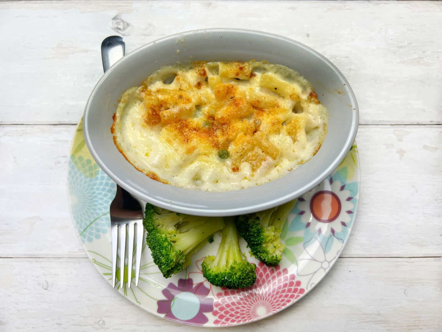 Macaroni cheese in a grey oval bowl served on a floral patterned plate with broccoli