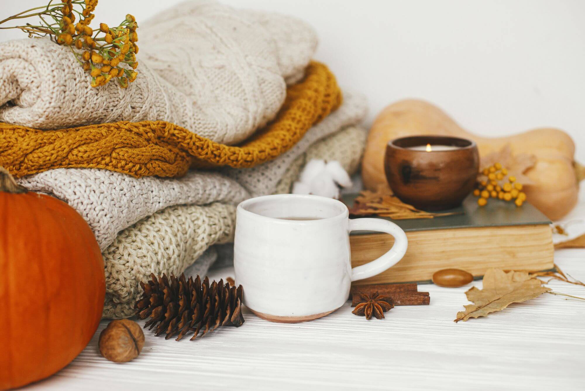 A white mug of tea with a stack of blankets behind and scattered autumn leaves and a pumpkin.