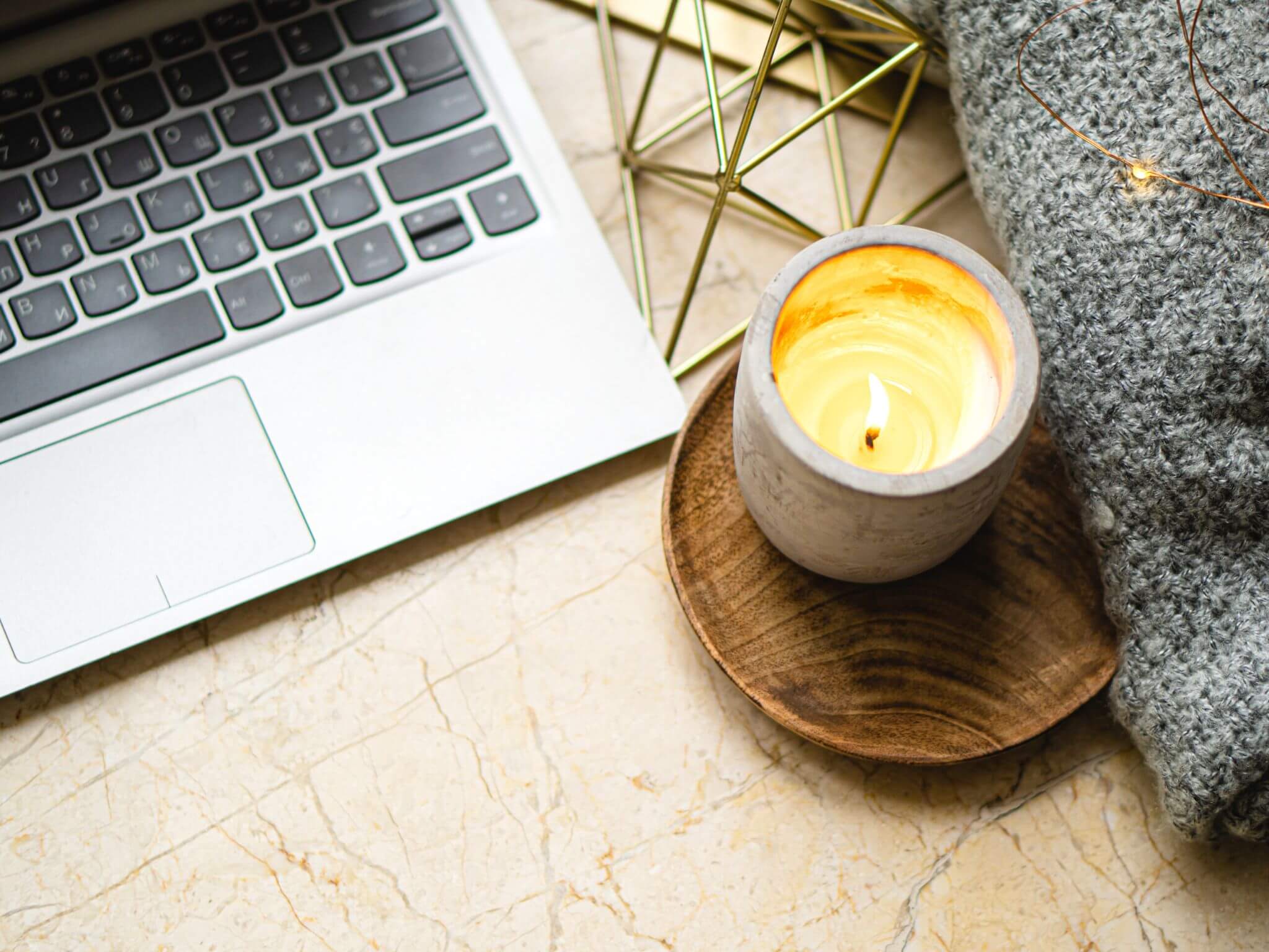 A open macbook pro on a desk top with a burning candle on a wooden coaster and a grey blanket to the side.
