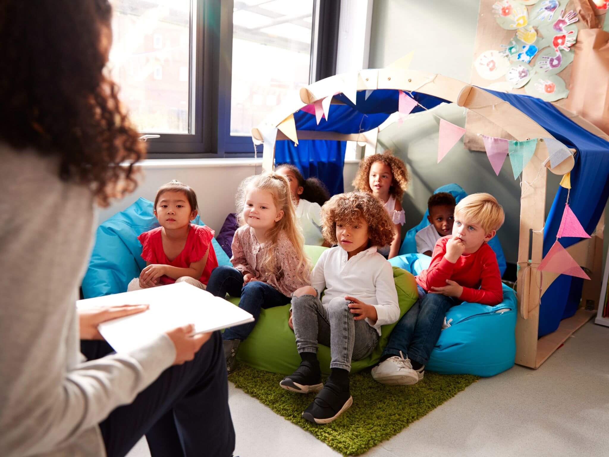 A class of infant aged children sitting on cushions listening to the teacher read a story.