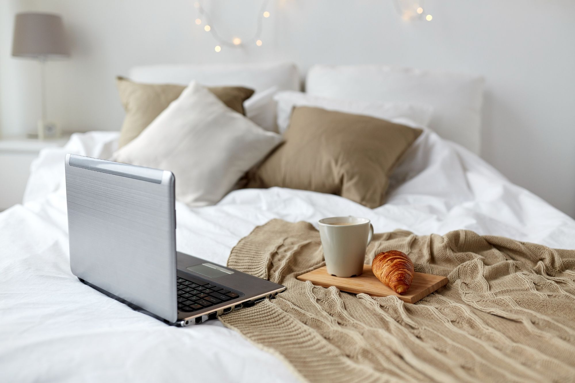 A bed with white sheets and a beige throw scattered across. There is also an open laptop on top of the bed along with a tray holding a mug of tea and a croissant. There are also fairy lights twinkling in the background.