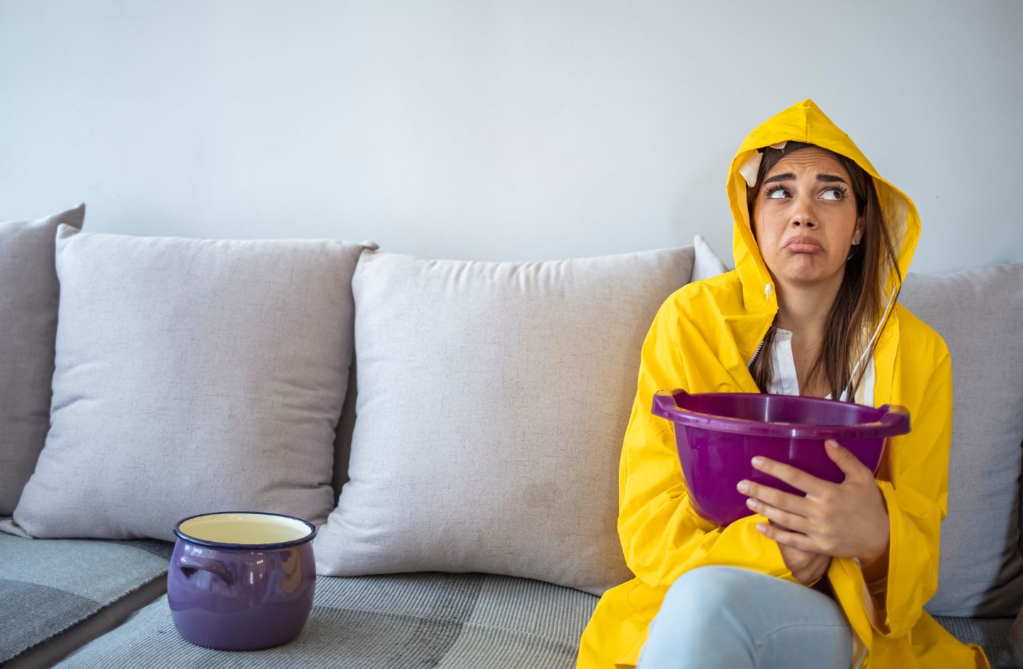 A lady wearing a yellow rain mac with hood, sitting on her sofa holding a bucket.