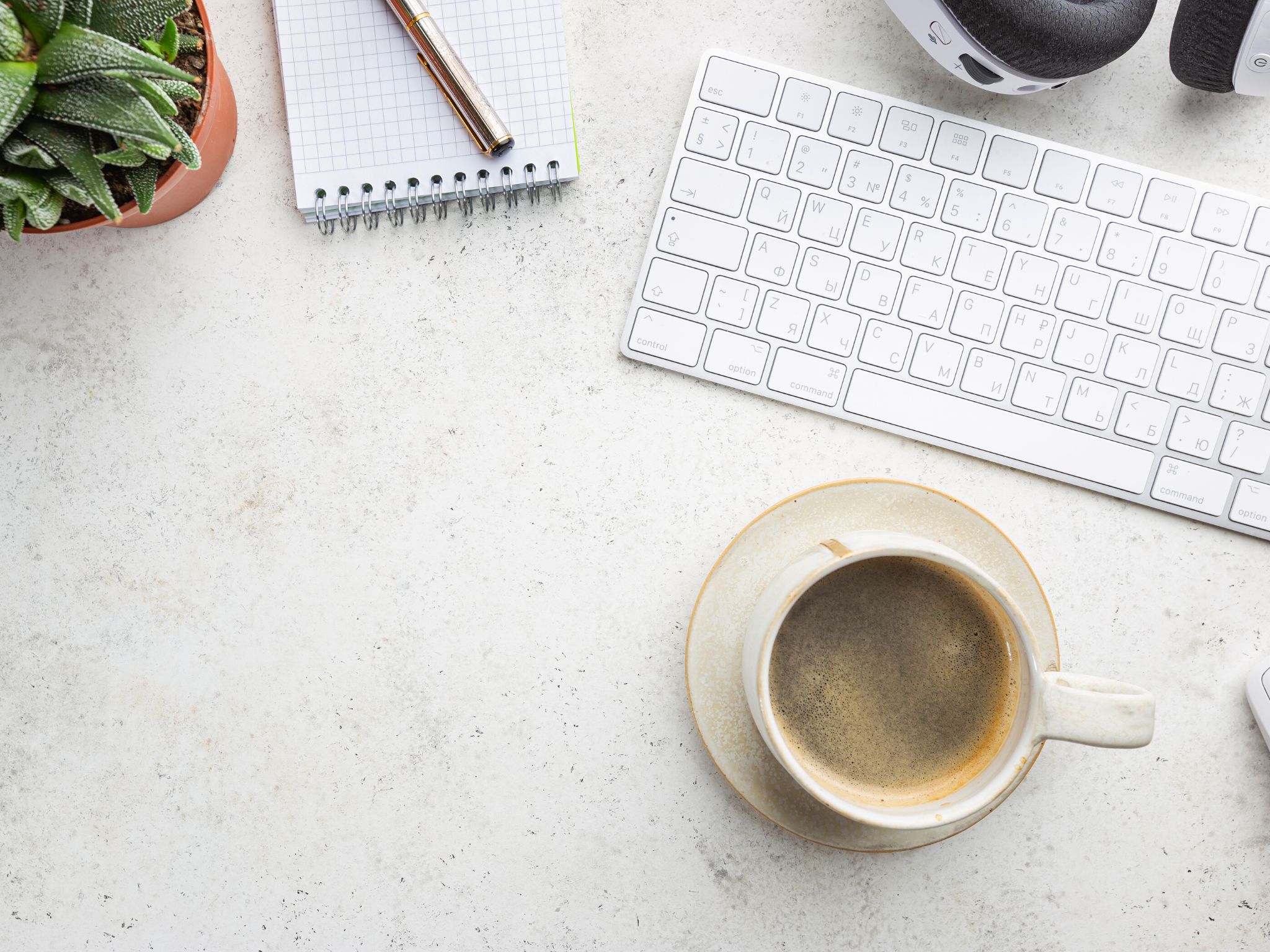A white desk top with a mug of coffee, an Apple keyboard, a notebook and pen, a cactus and a pair of white headphones.