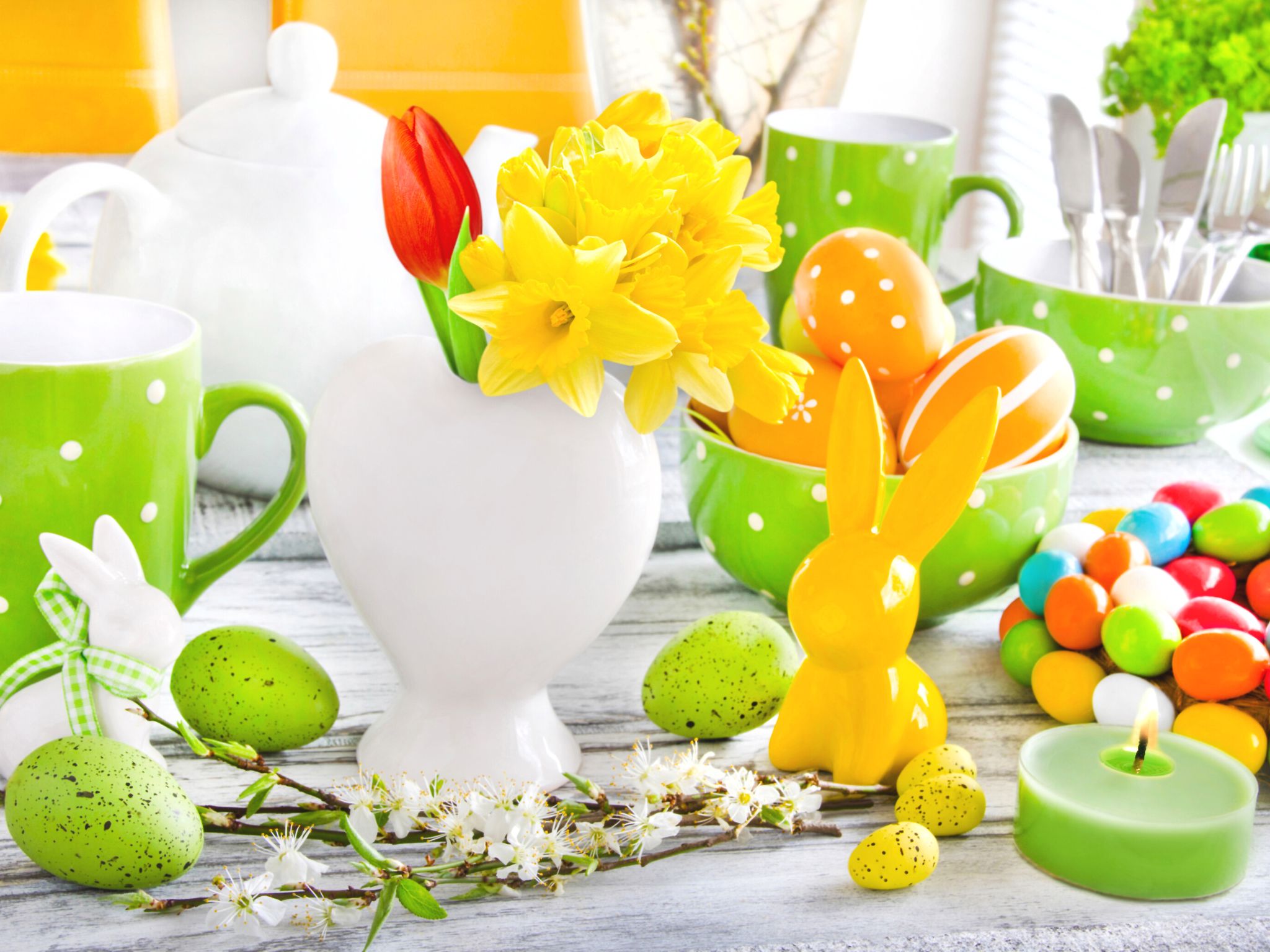 An Easter themed table with lime green mugs, bowls and eggs. There is also a white vase filled with daffodils and a ceramic yellow rabbit ornament by the side.