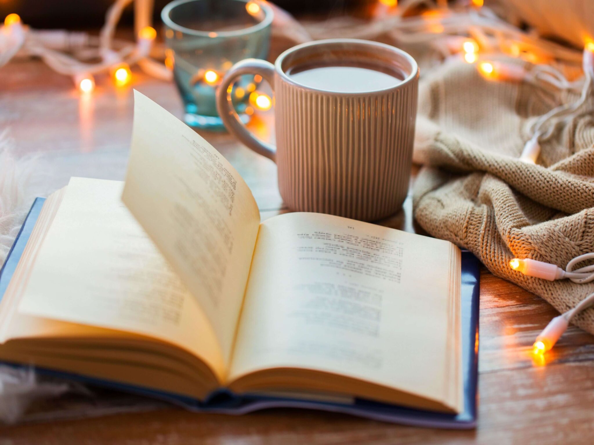 A coffee top with an open book on top. Just behind is a cup of coffee with a beige knitted blanket and warm white fairy lights.
