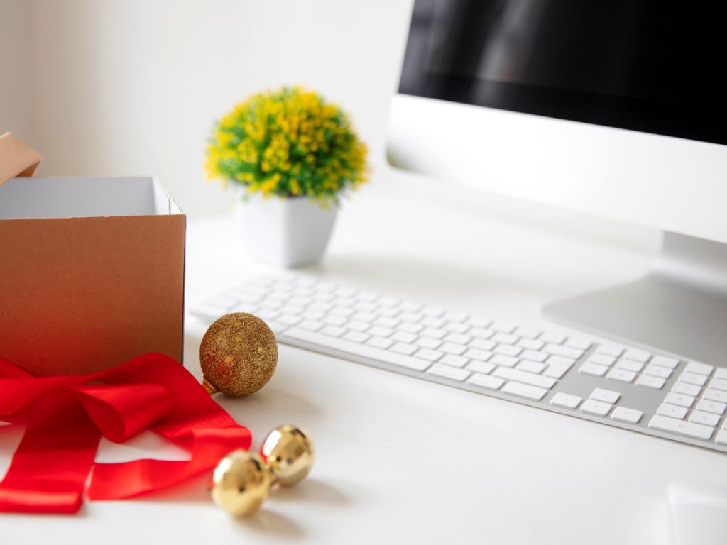 A white desk top with a MAC computer on top. There is also a an artificial green shrub in a white pot and a cardboard box with Christmassy red ribbon and gold baubles to the left.
