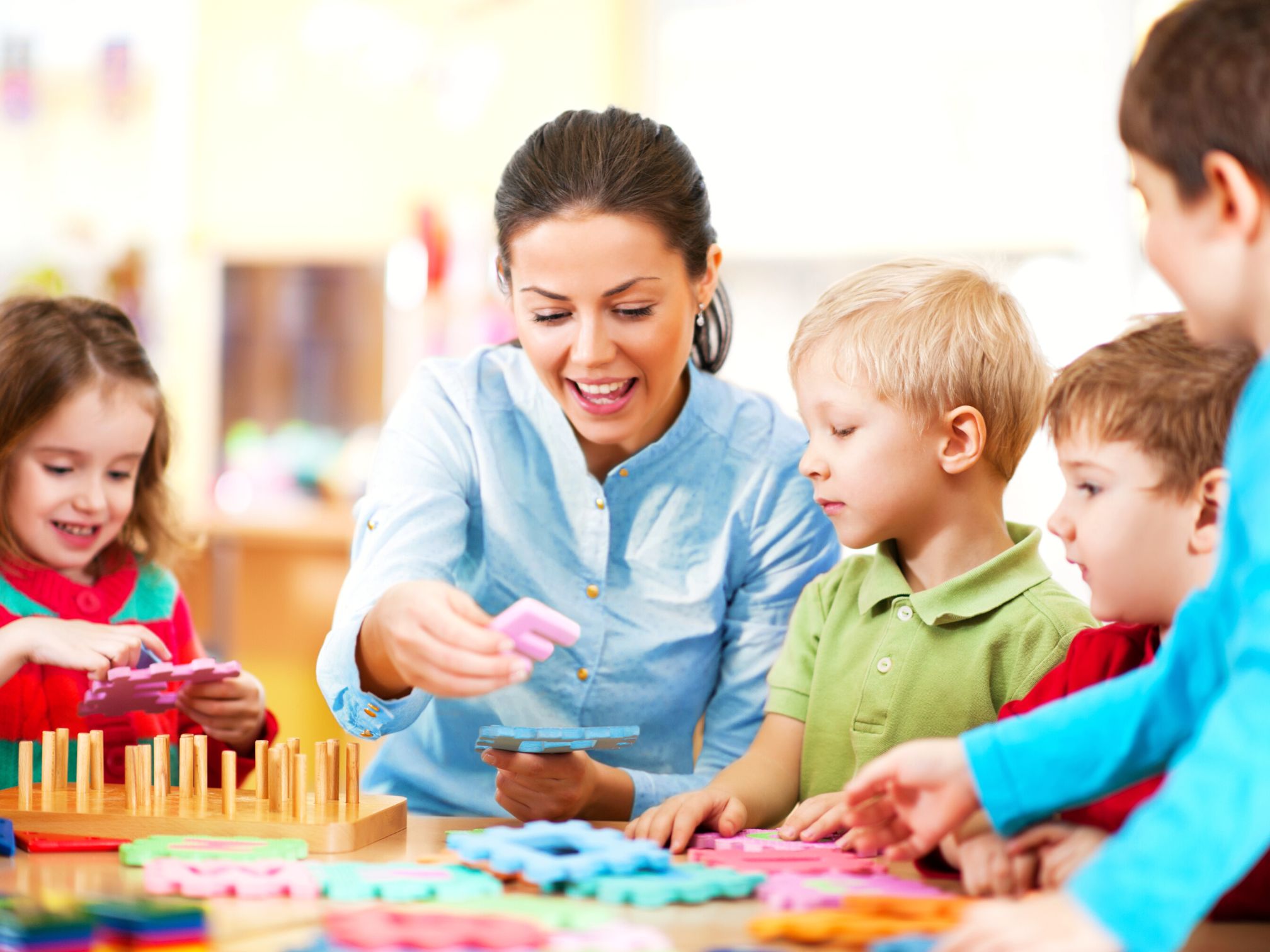 A nursery teacher sitting at a table with children, playing with puzzles.