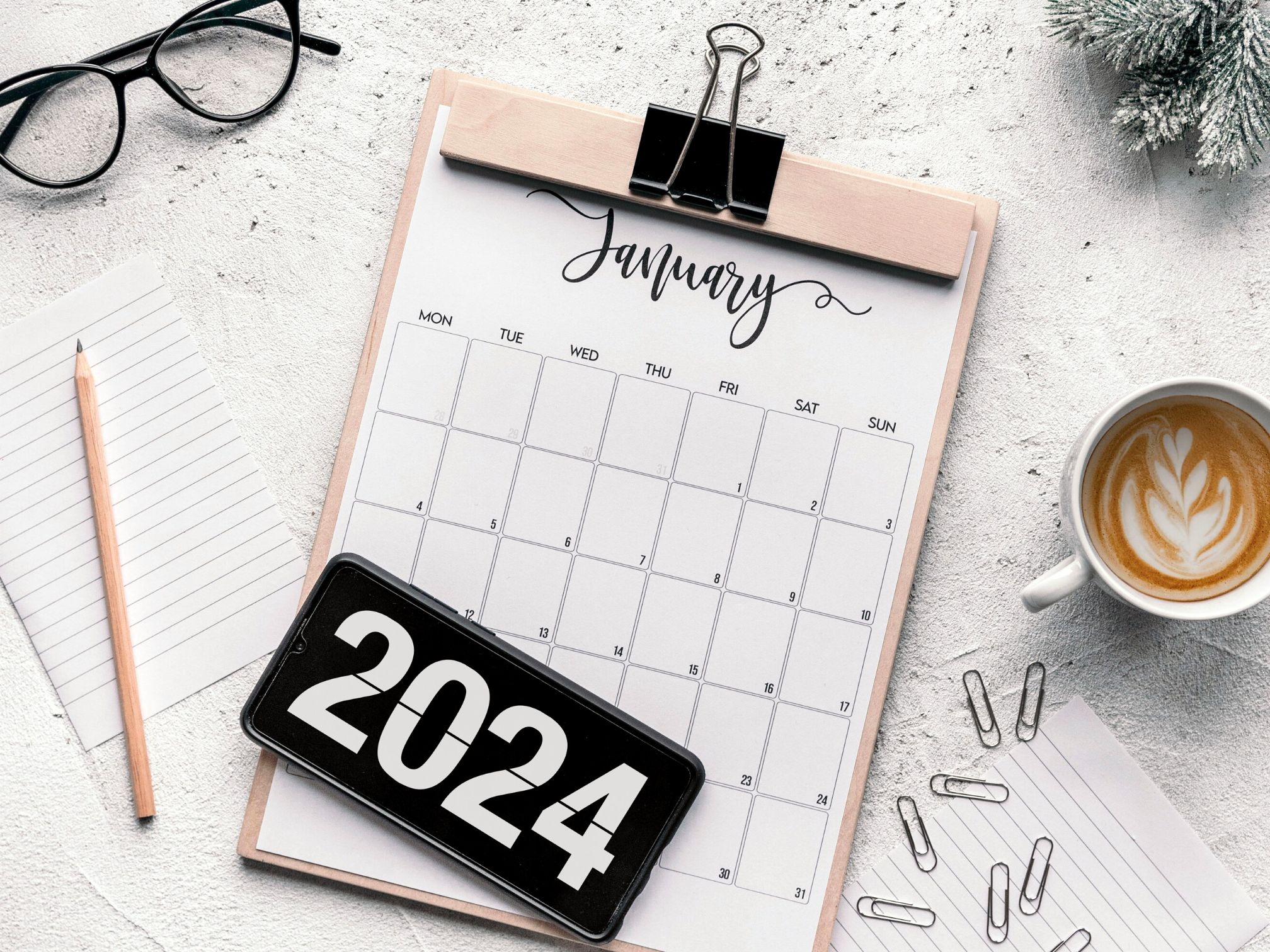 A white desk top with a clipboard and a a4 diary sheet attached to it. There is also a 2024 black sign along with a cup of tea and paper clips scattered around the cup. There is also a pair of black glasses and a pencil.