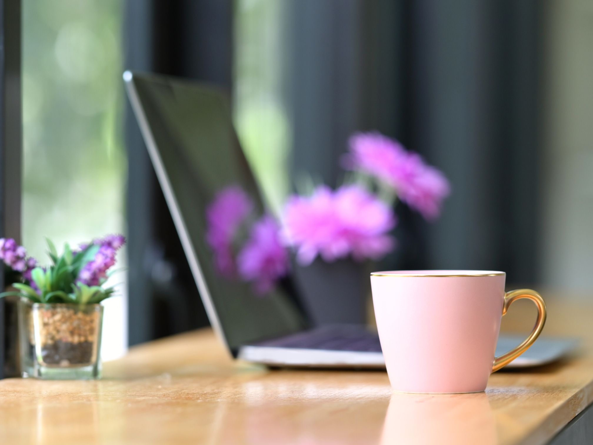 There is an open laptop on a beech worktop. By the side is a pink mug of tea and some pink asters in a vase next to it.
