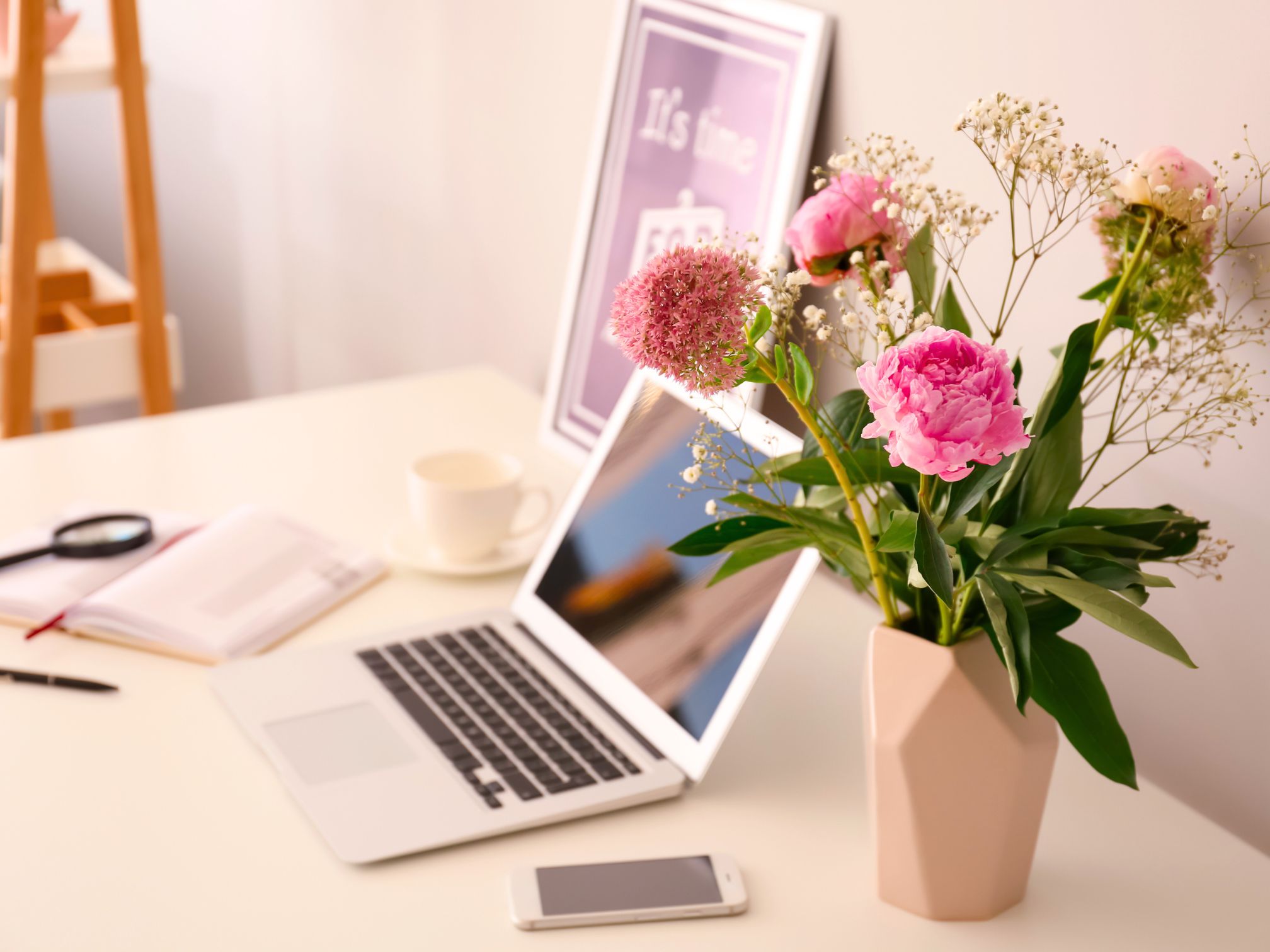 There is an open Macbook laptop on a white desk top. Beside this is a pastel pink vase holding pink cut roses. There is also a white iPhone, a white mug of tea and an open notebook and pen.