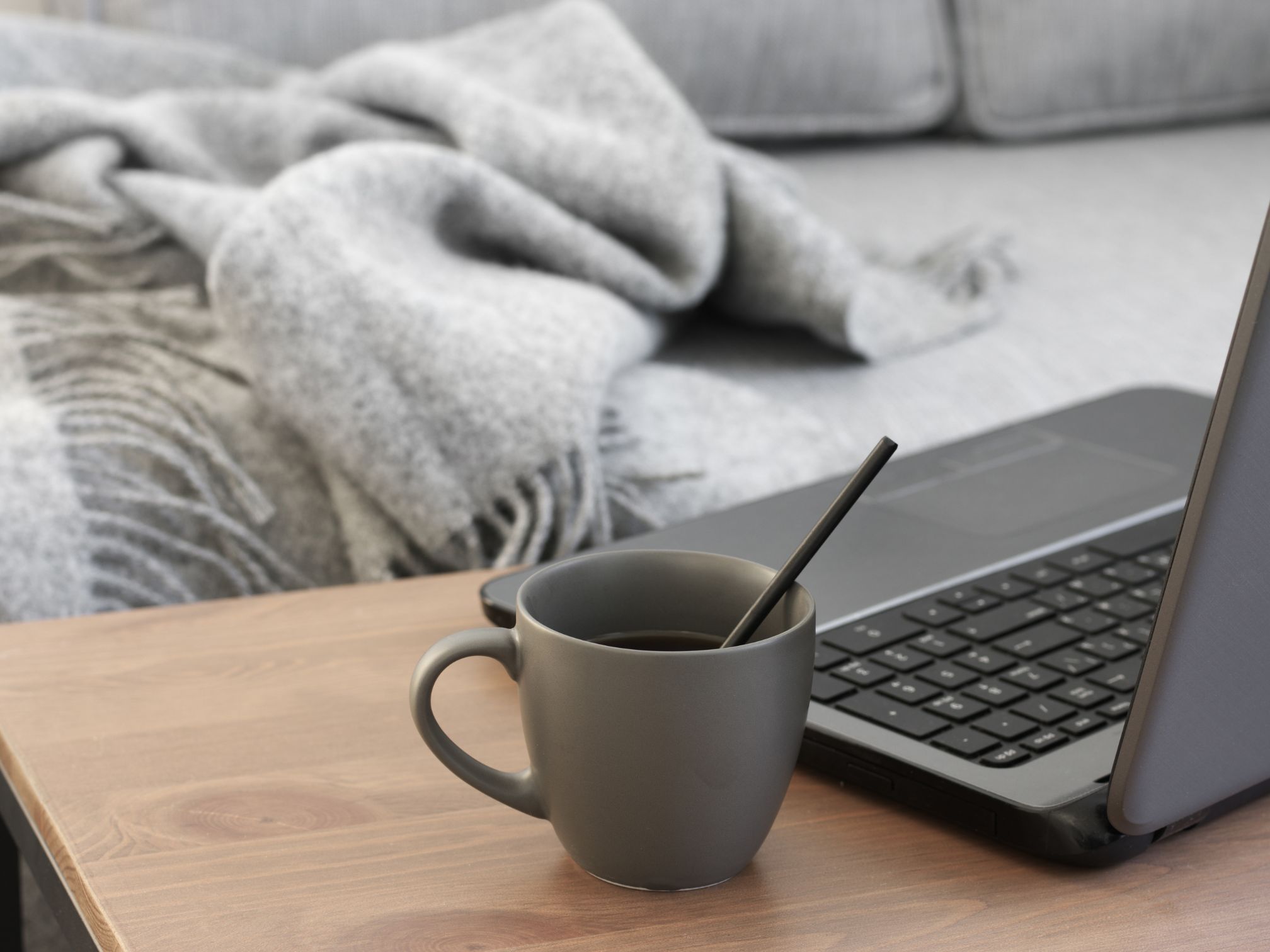 There is a grey soft fleece blanket draped on a light grey sofa. In front is a coffee table with an open macbook and a grey mug filled with coffee and a spoon inside.