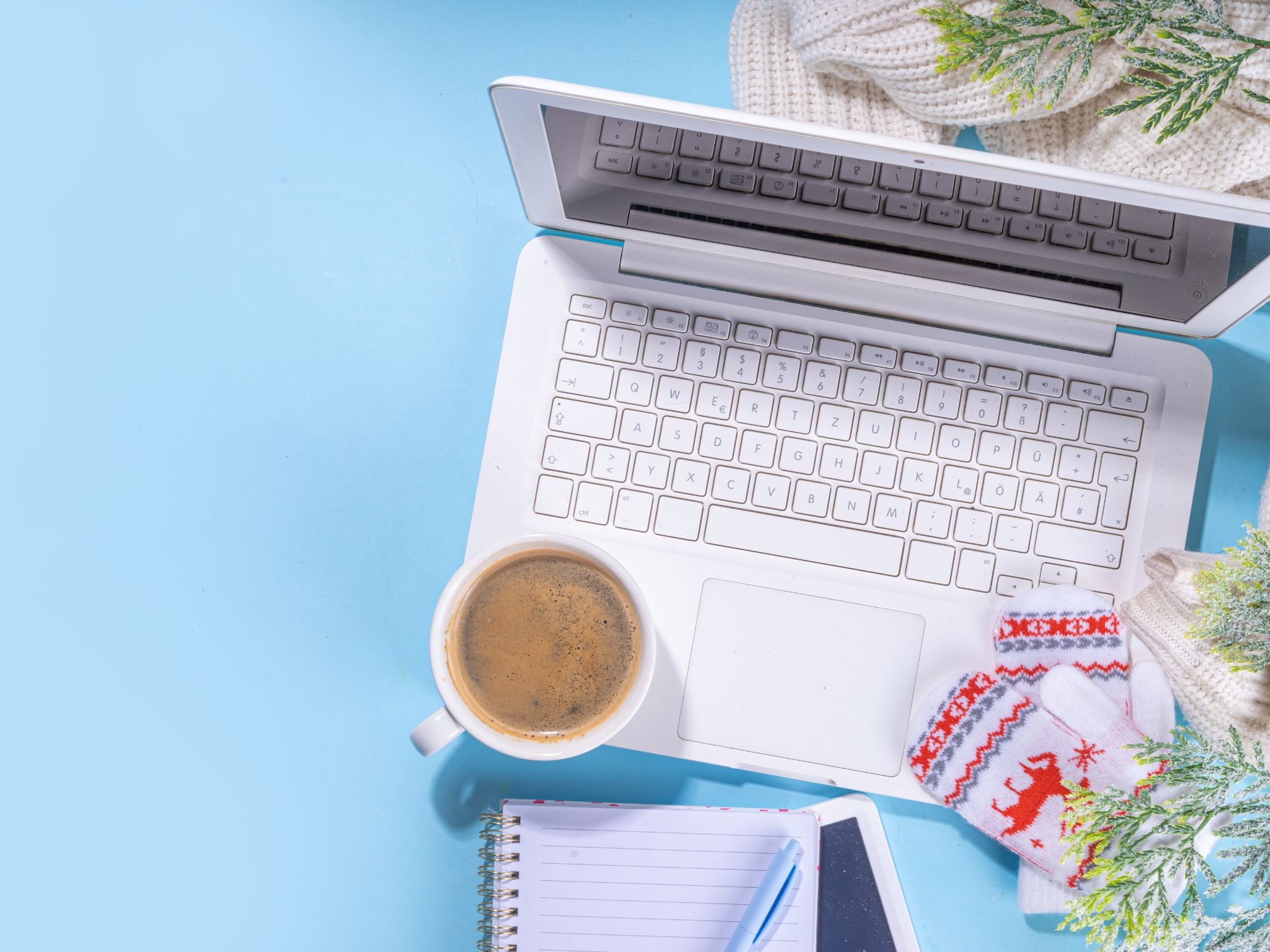 There's an open Macbook laptop on a blue desk top. There is also a frothy coffee in a white mug with a notebook to the side and a pair of white mittens with a reindeer pattern on. The desk is also decorated with spruce sprigs, giving it a festive vibe.