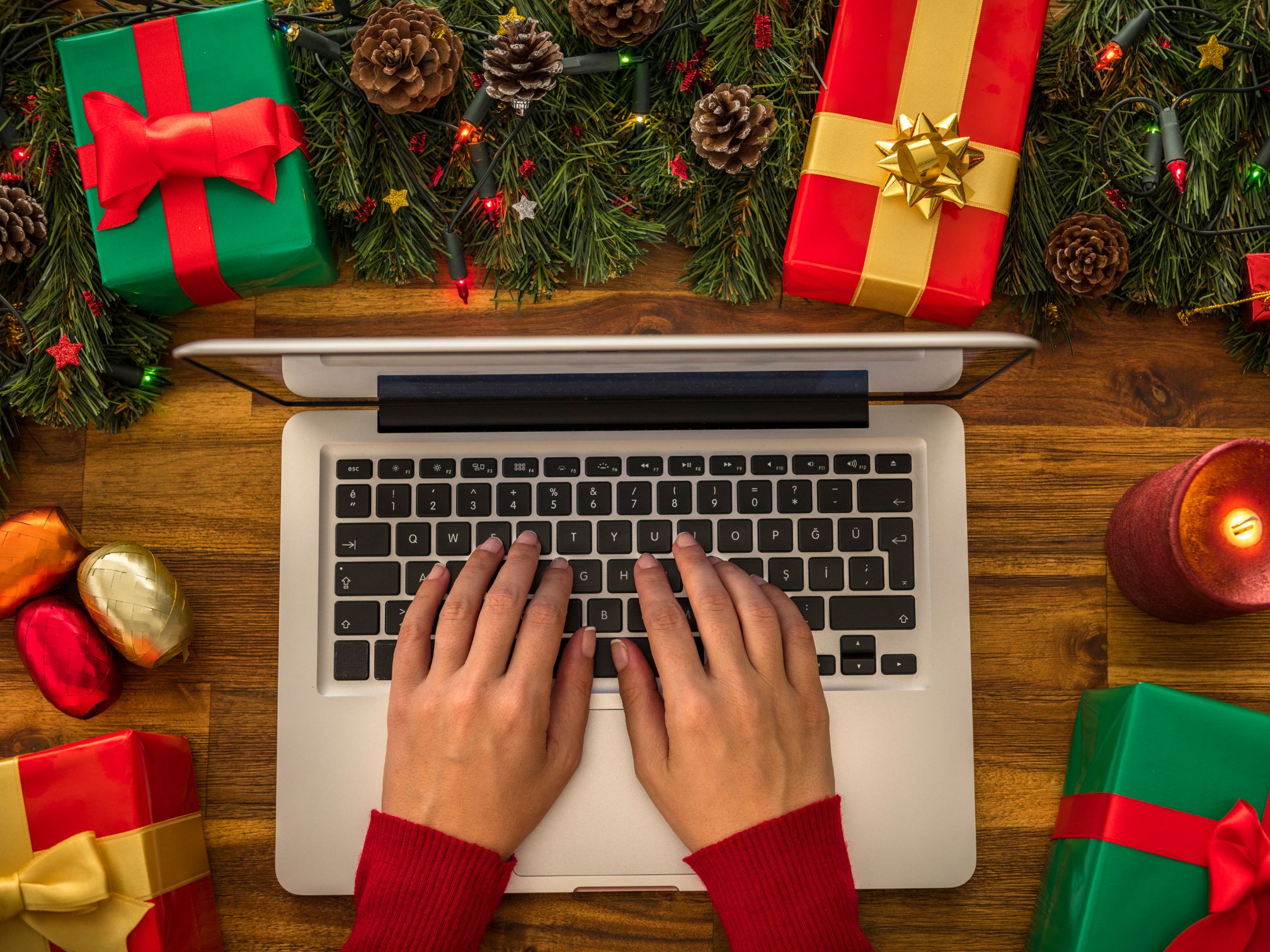 An open laptop on a desk with a lady's hands typing away. Scattered around the laptop is Christmas presents and Christmas tree foliage.