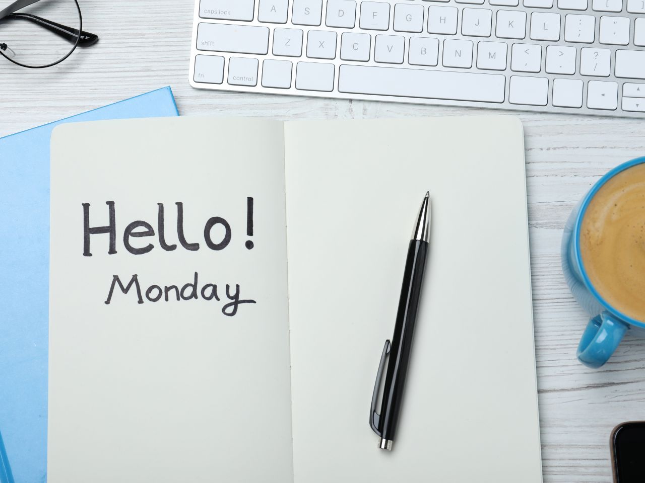 There is a white Apple keyboard on a white table top. In front is an open card with the words 'Hello Monday' written inside. There is a black pen on top and a mug of tea to the left.