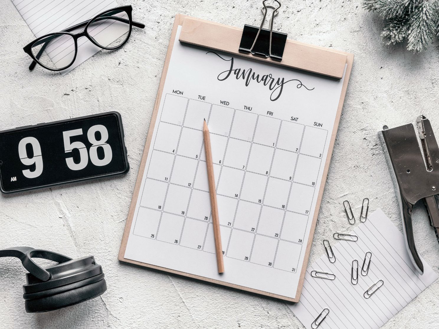On a white desk top there is clipboard with a calendar showing the month of January. To the side are scattered paper-clips, a pair of reading glasses and a clock.