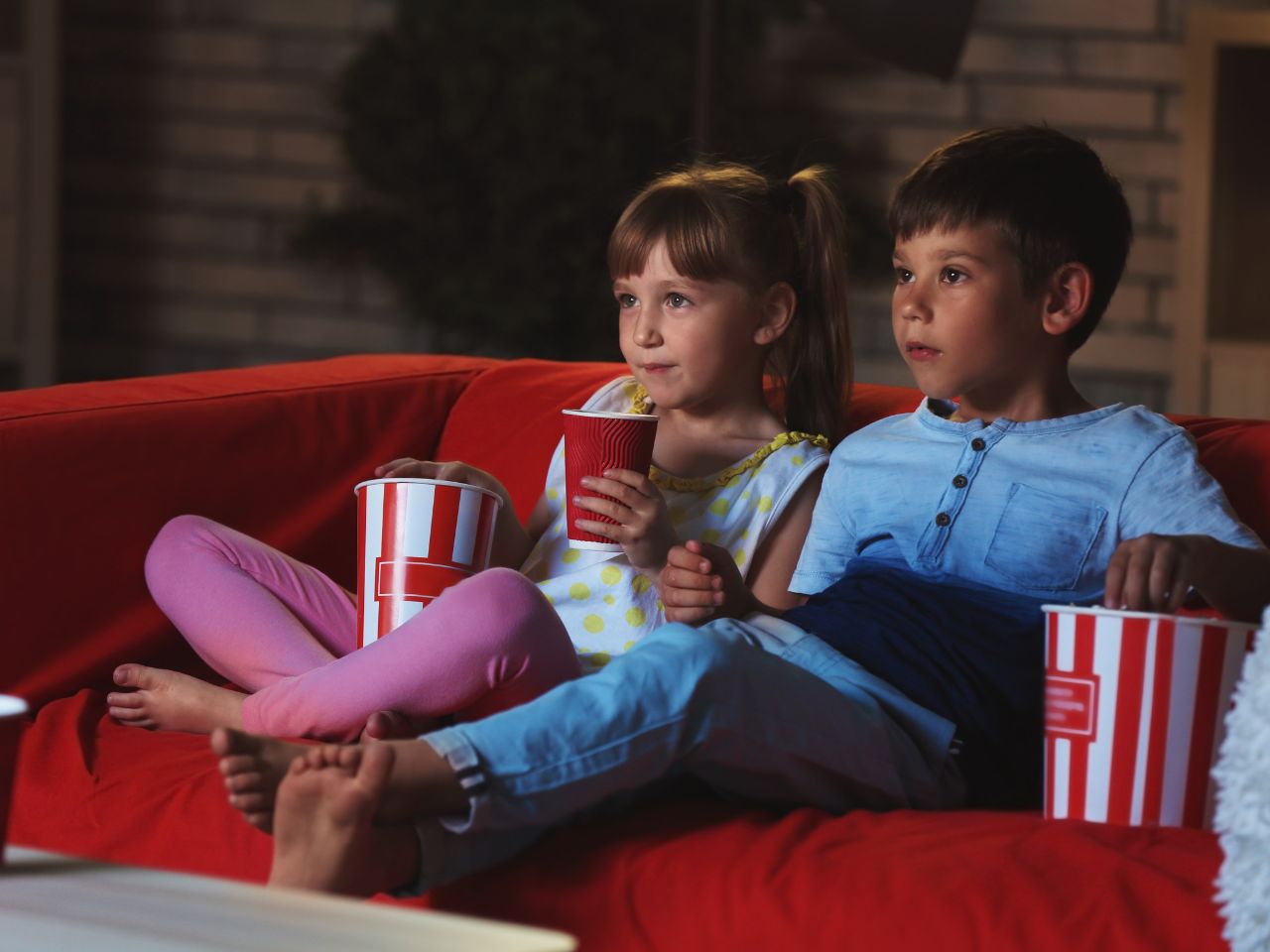 A young boy and girl (primary school age) sitting on a red sofa, watching TV with a tub of popcorn each and the little girl has a drink in a paper cup, enjoying a cosy Tv show together