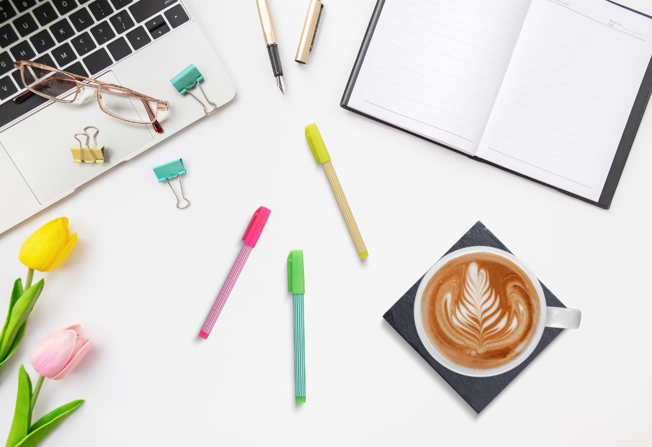 On a white desk top there is a macbook, an open blank notebook and three pink, yellow and green highlighter pens. There is also a white mug of coffee on a black slate coaster. To the left are two pretty pastel pink and yellow tulips.