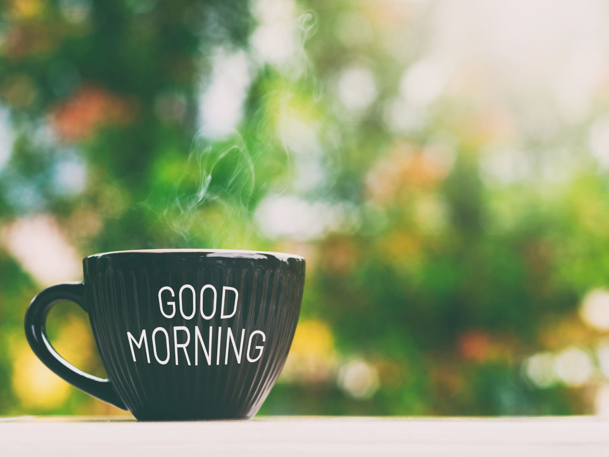 A black mug on a table outside. It has 'Good morning' on the front. You can see greenery blurred out in the background.