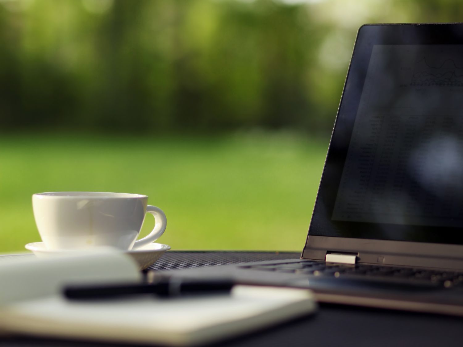 An open Macbook Pro on an outside picnic bench. By the side is a white cup filled with tea and an open diary and pen. The background is the garden.