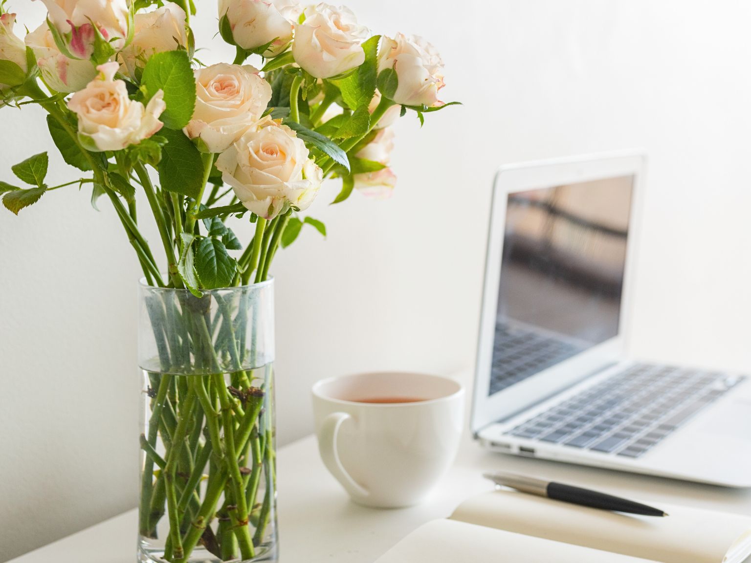 There is an open MacBook on the white desktop. To the left is a white mug filled with tea. A white notebook with a black pen and a clear glass vase holding several white roses.