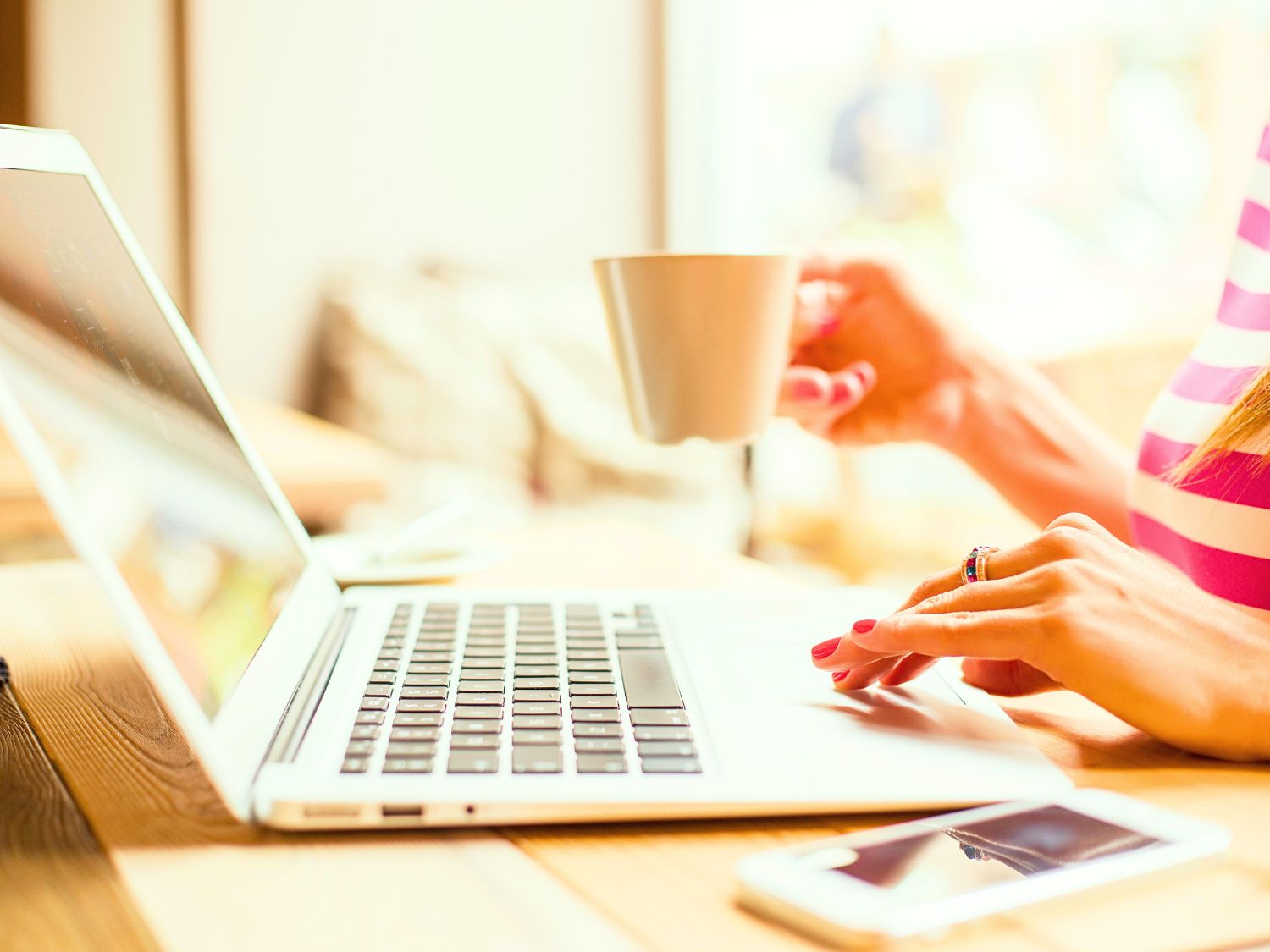 A close up of lady's arms sitting at her desk. She is typing with one hand on her laptop. The other hand is holding a white mug.