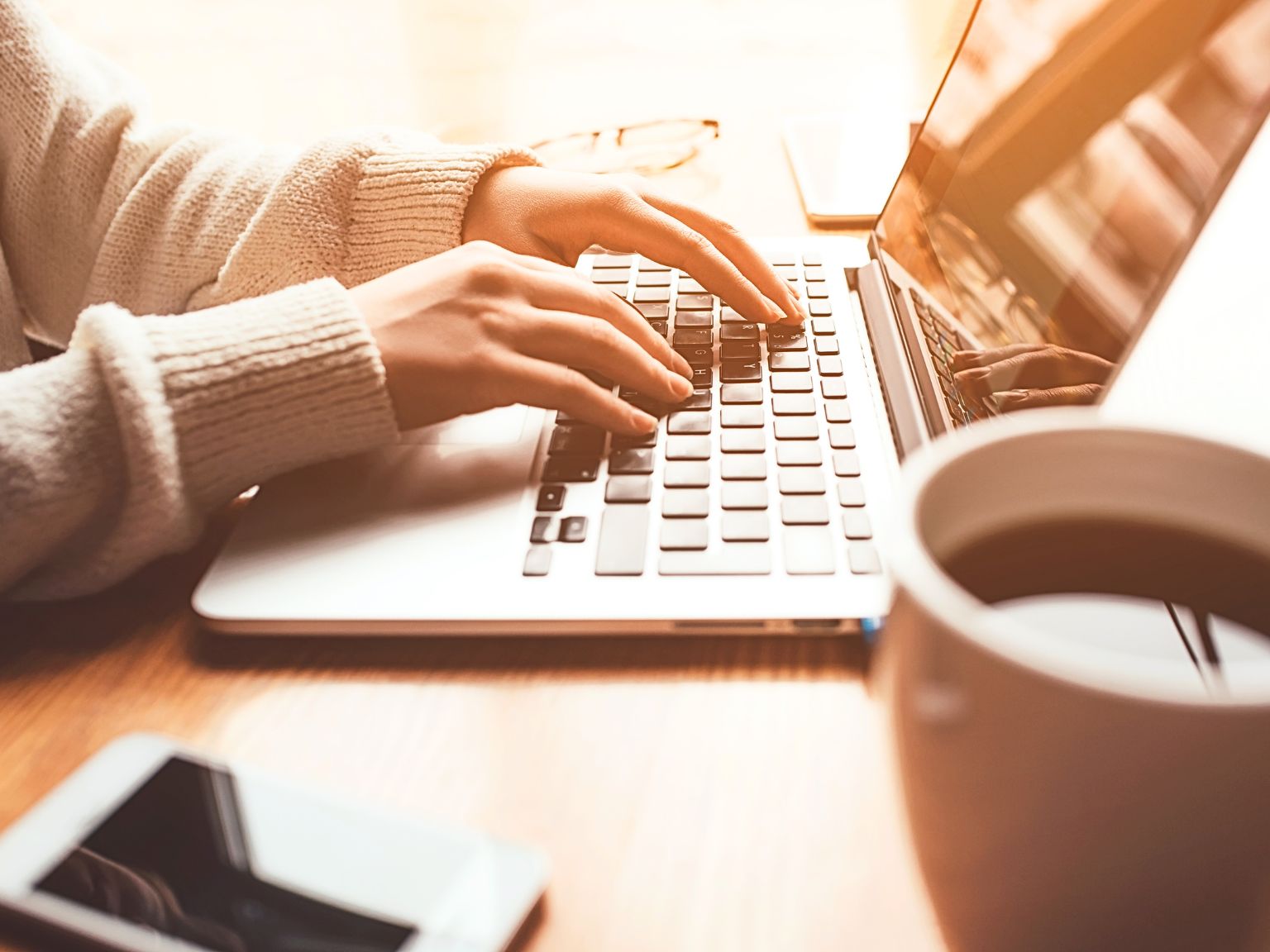 This is a close up of a ladies wrists and hands typing on an open MacBook. There is a black coffee to the side and an iPhone.