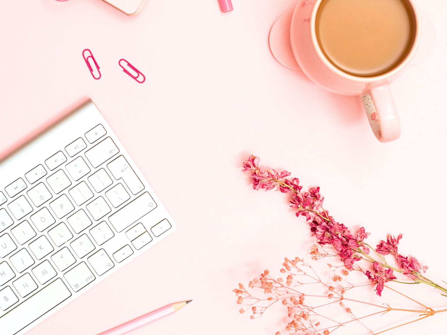On the top of a pink desk top, is a white Apple keyboard, some pink paper clips, a mug filled with tea and a sprig of pink dried flowers scattered below.