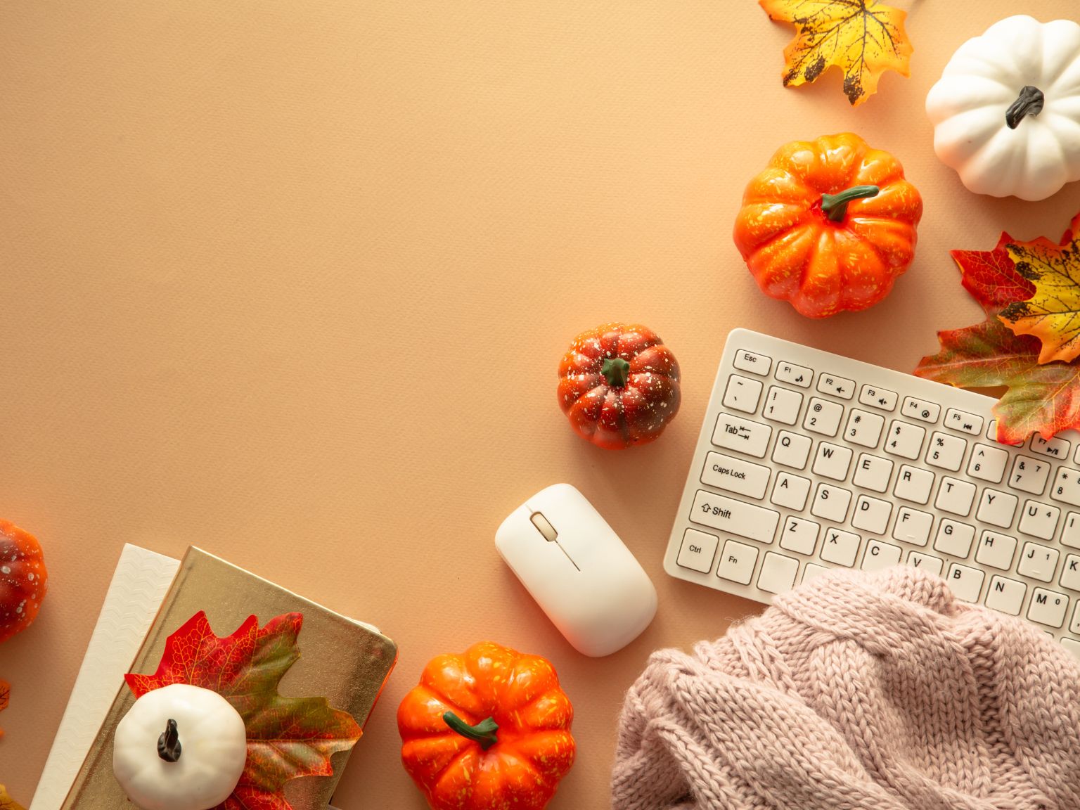 There is a white keyboard on a desk top surrounded by pumpkin ornaments, a blanket and a notebook.