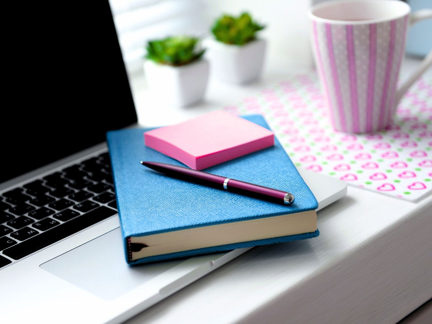 A laptop sits on a white desk with a blue notebook placed on top of it. A pen and pink sticky notes rest on the notebook. In the background, there is a patterned mug and two small potted plants, creating a calm, tidy workspace.