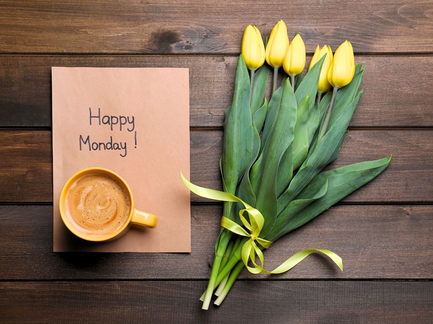 A piece of paper with the words 'Happy Monday' lies on top of a desk, with a bunch of yellow tulips on the right side.
