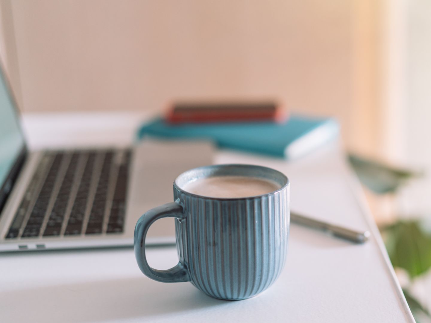 On a desk top, there is a blue mug filled with white tea. To the right is an open laptop, some stacked books and a pen.